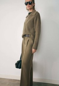 Suéter de canalé color caqui con cuello; pantalones beige de talle alto y con pinzas; bolso negro; grandes gafas de sol negras. Un atuendo simple y moderno.