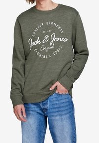 Sudadera verde con logo y texto impresos en blanco. Cuello redondo, puños acanalados, ajuste informal, combinada con vaqueros azules. Textura de tela suave.