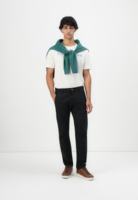 Junger Mann steht mit Händen in den Taschen, trägt ein weißes Grafik-T-Shirt, schwarze Hose, braune Sneakers und einen grünen Pullover um die Schultern gebunden.