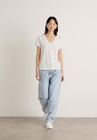 Vit t-shirt med v-hals i bomull matchad med ljusblå, löst sittande jeans, bärs med vita sportskor mot en neutral bakgrund.