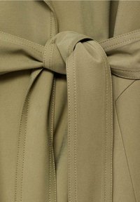 Krawat z tkaniny oliwkowozielonej z wiązaniem w supeł, wykonany z elastycznego materiału. Tekstura jest gładka, z widocznym przeszyciem wzdłuż krawędzi.