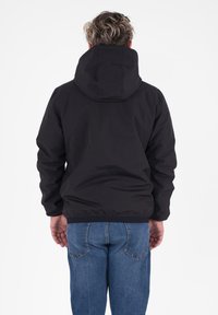 Persoon gezien van achteren, gekleed in een zwarte capuchonjack en blauwe spijkerbroek, tegen een eenvoudige witte achtergrond.