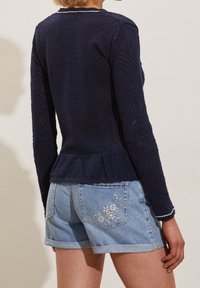 Námořnický pletený top s texturovaným vzorem, dlouhými rukávy a peplum lemem. Nosit s světle modrými džínovými šortkami s květinovou výšivkou.
