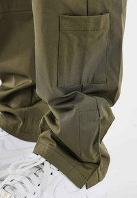 Olijfgroene cargobroek met een gladde textuur, voorzien van een zijzak en elastische cuffs, gedragen met witte sneakers.