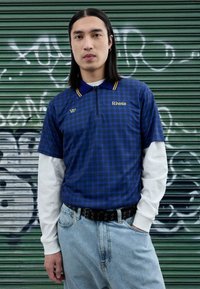 Plava polo majica na kockice s polu-rajfešlus ovratnikom i žutim detaljima, nošena preko bijele majice s dugim rukavima, u kombinaciji sa svijetloplavim trapericama i crnim remenom.