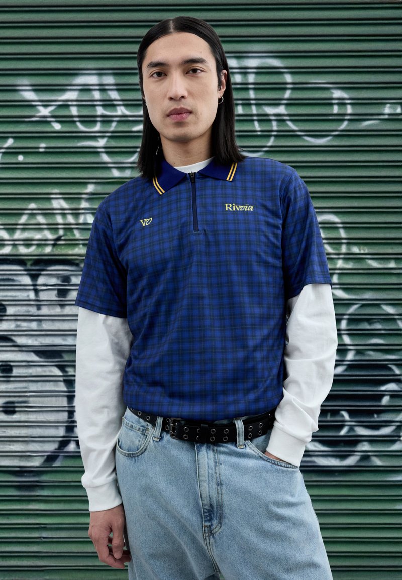 Plava polo majica na kockice s polu-rajfešlus ovratnikom i žutim detaljima, nošena preko bijele majice s dugim rukavima, u kombinaciji sa svijetloplavim trapericama i crnim remenom.