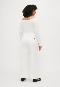 Hvit langermet topp med off-shoulder design, kombinert med hvite vide bukser og svarte flate sko. Slank tekstur og minimalistisk stil.