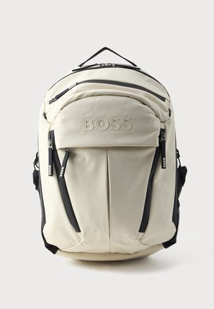 Lys beige ryggsekk med svarte detaljer, som har flere glidelåslommer, ergonomiske stropper og et hevet logo på forsiden.