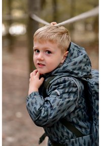 Grå mønstret vinterjakke med hætte, med grønne detaljer. Barnet holder i en rem, med en rygsæk synlig på ryggen.