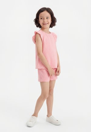 Mladé dievča sa usmieva, nosí ružový textúrovaný bezrukávový top a zodpovedajúce šortky s bielymi teniskami, stojí na pozadí bielej steny.