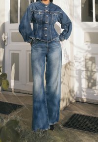 Jeansjacka med hög krage och puffärmar, tillsammans med vida blå jeans. Båda plagg har en urtvättad känsla och klassiska designinslag.