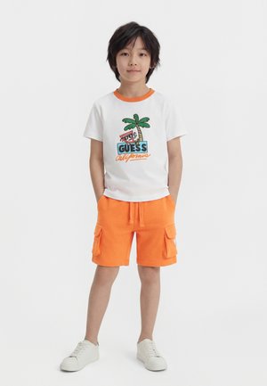 Αγόρι που στέκεται με τα χέρια στις τσέπες, φοράει λευκό μπλουζάκι με γραφικό φοίνικα και πορτοκαλί cargo σορτς με λευκά αθλητικά παπούτσια.