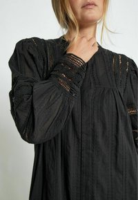 Black blouse featuring intricate lace accents on the sleeves, textured fabric, and vertical stripes, with a relaxed fit and button detail.