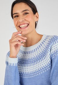 Pull en tricot bleu clair avec un col rond, présentant un motif géométrique blanc au niveau du devant et des poignets côtelés. Texture douce.