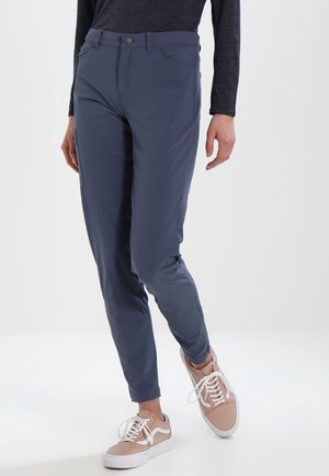 Person trägt eng anliegende marineblaue Hose und hellrosa Turnschuhe, steht vor einem weißen Hintergrund mit entspannten Händen seitlich am Körper.