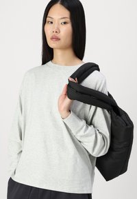 Šedá dlouhá tričko s kruhovým výstřihem, doplněné černou polstrovanou taškou nošenou přes rameno. Hladká textura a volný střih.