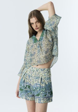 Jonge vrouw draagt een losse top met een bloemen paisley patroon en bijpassende shorts met hoge taille, poseert met één arm omhoog boven haar hoofd.