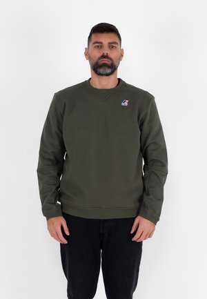 Felpa verde oliva con scollo rotondo, polsini e orlo a coste; presenta un piccolo logo sul petto. Indossata con jeans scuri.