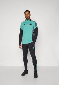Man standing wearing teal and black Adidas sportswear with logo and three stripes, black athletic shoes, against plain background.