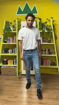 Fehér, kerek nyakú póló, világoskék farmer enyhe kopott hatással, fekete cipő, beltéri környezet sárga falakkal és zöld polcokkal.