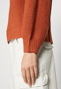 Rustykalny pomarańczowy sweter z dzianiny z teksturowanym wzorem i ściągaczami, w połączeniu z jasnymi spodniami. Zbliżenie na rękaw.