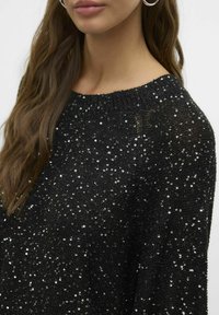 Pull noir avec un design de tricot texturé, orné de sequins argentés, encolure ronde et poignets côtelés, partiellement visible.