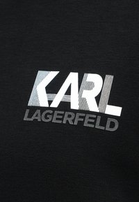 Λευκό "KARL" με ριγέ μοτίβο πάνω από το γκρι λογότυπο "LAGERFELD" τυπωμένο πάνω σε μαύρο υφασμάτινο φόντο.