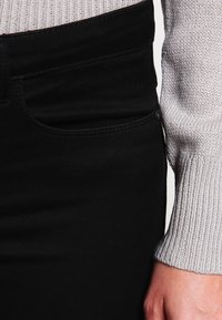 Pantalones vaqueros de mezclilla negra con una textura suave, que presentan un bolsillo clásico y detalle de botón de metal, combinados con un suéter de rayas gris claro.