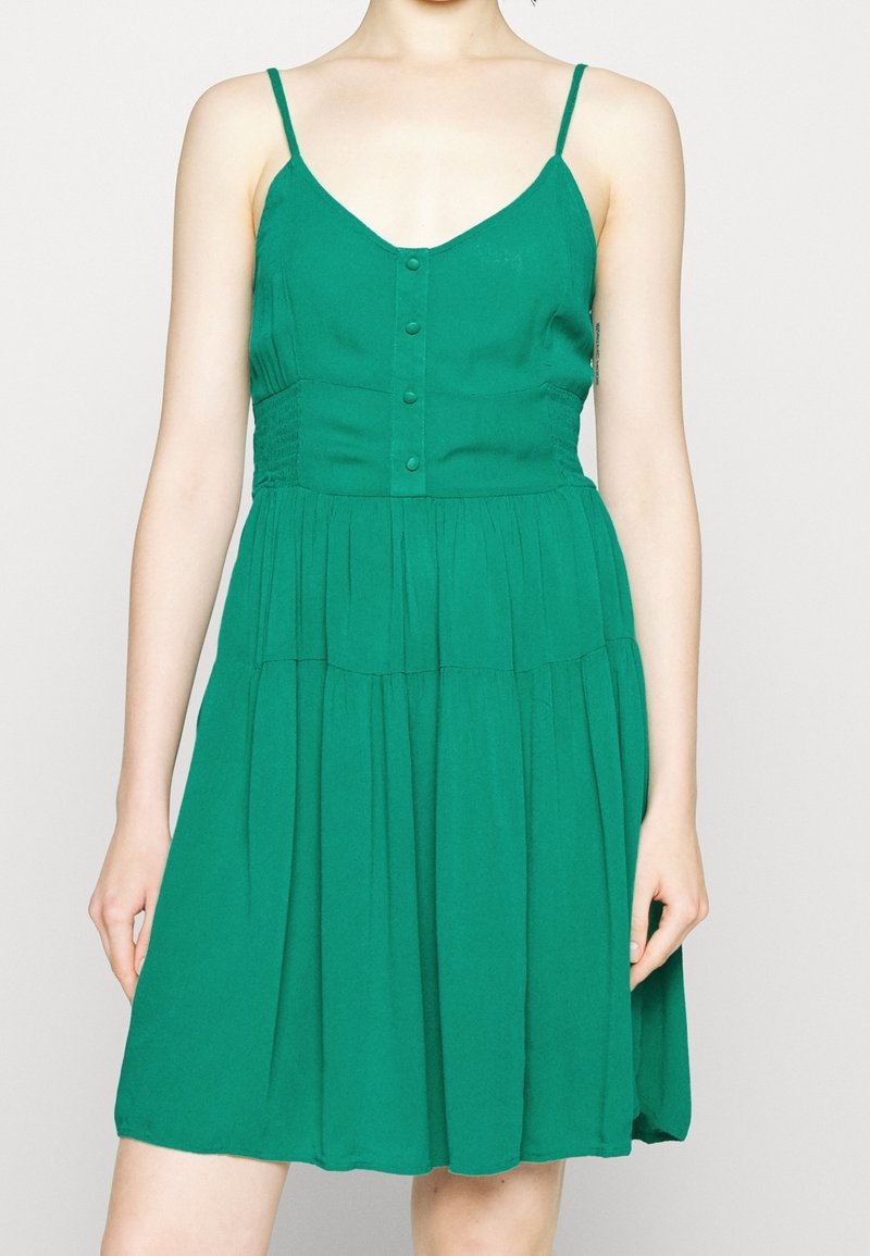 Femme portant une robe verte sans manches avec des boutons sur le devant et une jupe froncée, debout devant un fond uni.