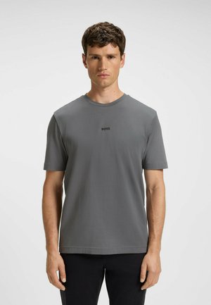 Man med kort brunt hår som bär en grå BOSS t-shirt och svarta byxor, stående mot en enkel vit bakgrund.