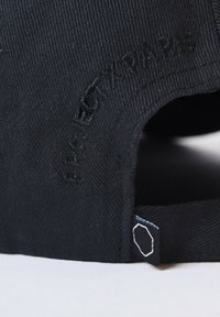 Gorra de algodón negra con tejido texturizado, logotipo bordado y una pequeña etiqueta con un diseño geométrico blanco en el costado.