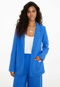 Modrá sako vyrobené z ľahkej tkaniny s klopovým golierom, dvoma prednými vreckami a ružovým akcentovým štítkom. Nosené cez biele tričko a zodpovedajúce nohavice.