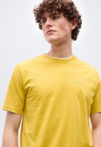 Νεαρός άνδρας με σγουρά μαλλιά που φοράει ένα κίτρινο κοντομάνικο T-shirt BOSS, κοιτάζοντας ελαφρώς προς τα πλάγια μπροστά σε απλό φωτεινό φόντο.