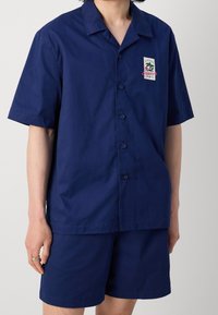 Mörkblå kortärmad skjorta med knäppning och avslappnad passform, med en bröstficka, i kombination med matchande shorts, tillverkad av ett lätt material.