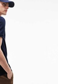 Camisa polo azul marino de manga corta, junto a pantalones marrones. El modelo está de pie con las manos en los bolsillos, vistiendo una gorra a juego. Fondo blanco.