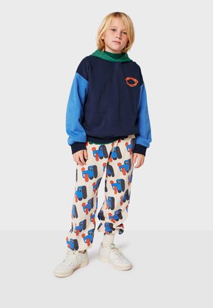 Navy and light blue color-blocked sweatshirt with green hood; cream pants featuring colorful cartoon designs; white sneakers.