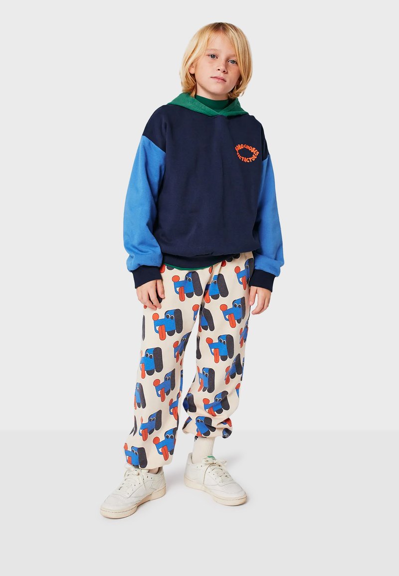 Navy- und hellblau gestreifter Sweatshirt mit grüner Kapuze; cremefarbene Hose mit bunten Cartoon-Mustern; weiße Sneakers.