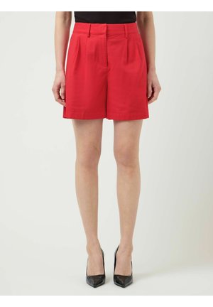 Vrouw draagt rode plissé shorts met hoge taille, een zwarte top en zwarte puntige hoge hakken, staand tegen een effen achtergrond.