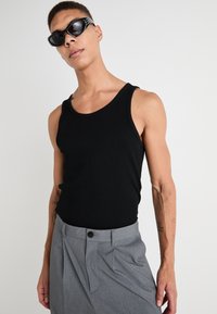 Débardeur noir côtelé avec larges bretelles, associé à un pantalon gris ajusté avec un détail de bouton. Le modèle a les cheveux courts et porte des lunettes de soleil.