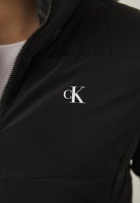 Sort pufferjakke med lynlåsfortil, der har et hvidt "cK" logo på venstre side. Glat tekstur med et slankt, tætsiddende design.