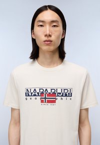 Kortærmet creme t-shirt med et blåt og rødt norsk flaggrafik, med teksten "NAPAPIJRI" og "geographic" i marineblå.