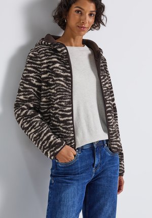 Femme portant une veste en polaire zèbre brun et blanc par-dessus un pull léger avec un jean bleu, se tenant avec une main dans sa poche.