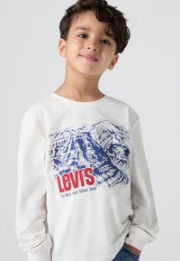 Dugi rukav bijela majica s plavim grafikom planine i crvenim 'Levi's' logotipom. Pamuk, ležerni kroj, okrugli izrez i rebrasti manžetni.