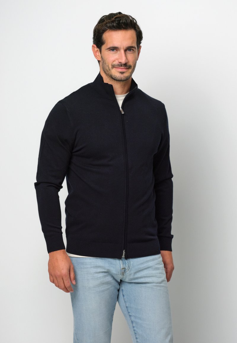 Maglione zip con collo alto e maniche lunghe di colore blu navy, realizzato in un materiale morbido. Dettagli notevoli includono una zip argentata e polsini a costine.
