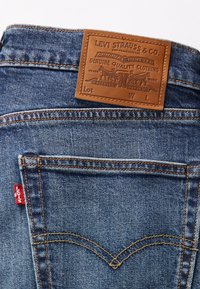 Zilas denima bikses ar brūnu ādas etiķeti, uz kuras ir rakstīts "Levi Strauss & Co." un oranžām šuvēm. Aizmugurējā kabatā ir sarkana logo etiķete.