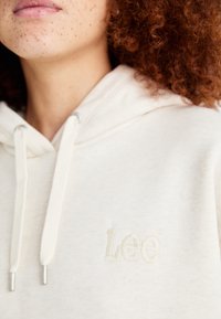 Gros plan du cou et du bas du visage avec des taches de rousseur et des cheveux roux bouclés portant un sweat à capuche blanc avec "Lee" brodé sur la poitrine.