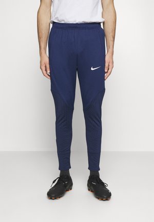 Homme portant un pantalon de sport Nike bleu marine et des crampons noirs, debout devant un fond clair uni.