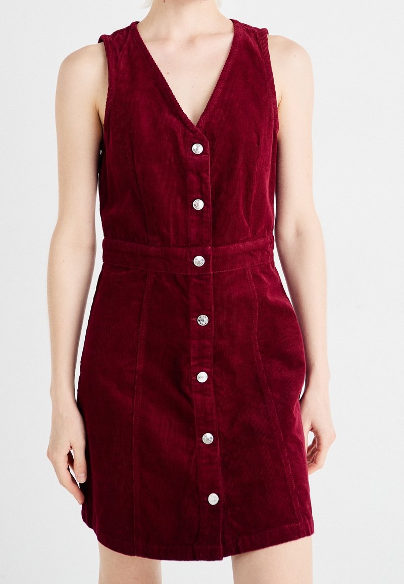 Robe bordeaux en velours côtelé sans manches avec des boutons argentés sur le devant et une taille ajustée, portée par une personne sur un fond uni.