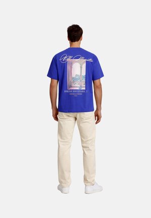 Man wearing blue graphic t-shirt featuring an archway and balcony print, beige pants, and white sneakers, standing facing away.