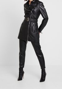 Svart läder trench coat med bälte, dragkedja, sidofickor och axelremmar, ihopkopplad med svarta snäva byxor och klackade snörstövlar.